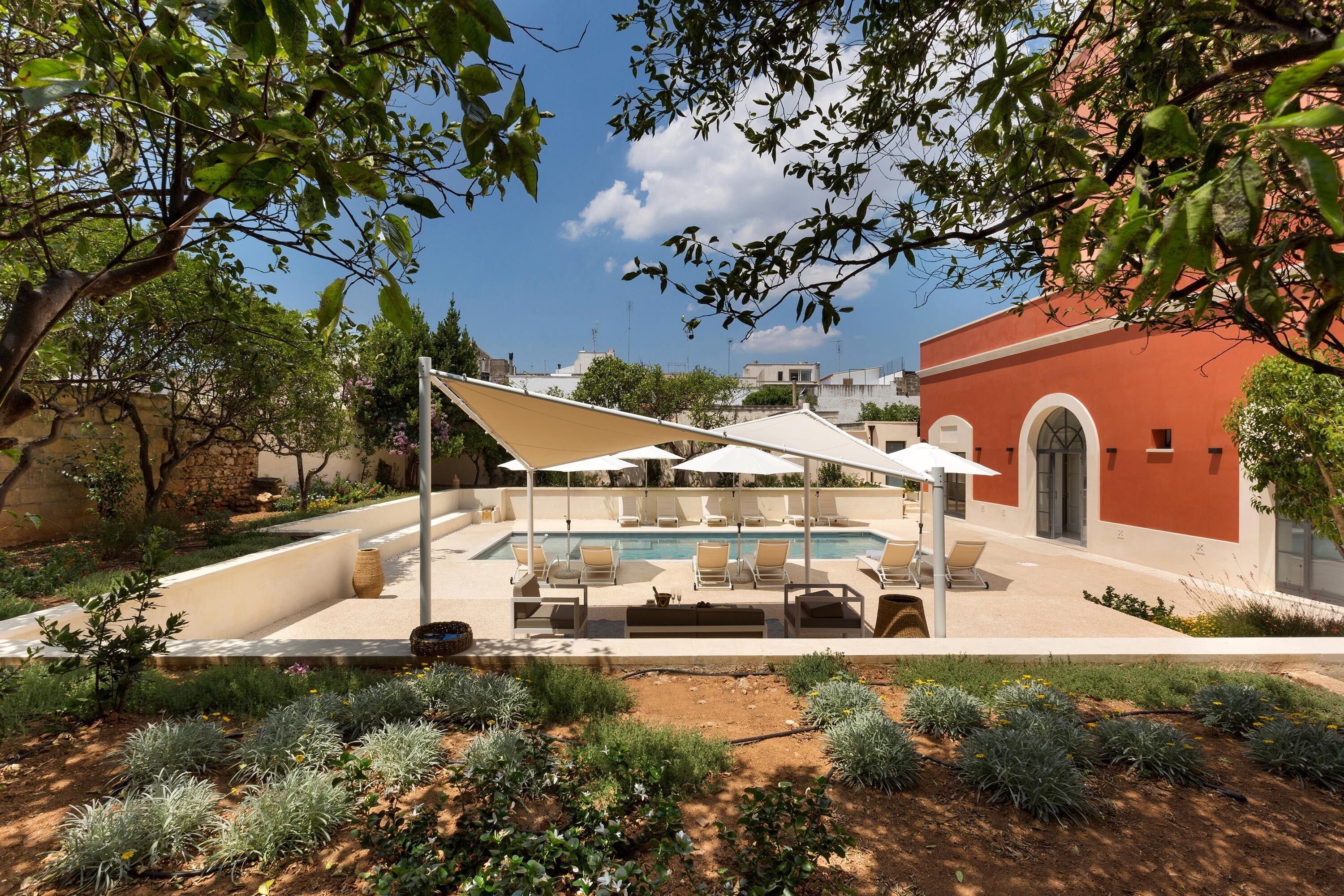 A pool area with lounge chairs and umbrellas, surrounded by landscaped gardens and a red building in the background.