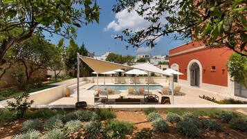 A pool area with lounge chairs and umbrellas, surrounded by landscaped gardens and a red building in the background.