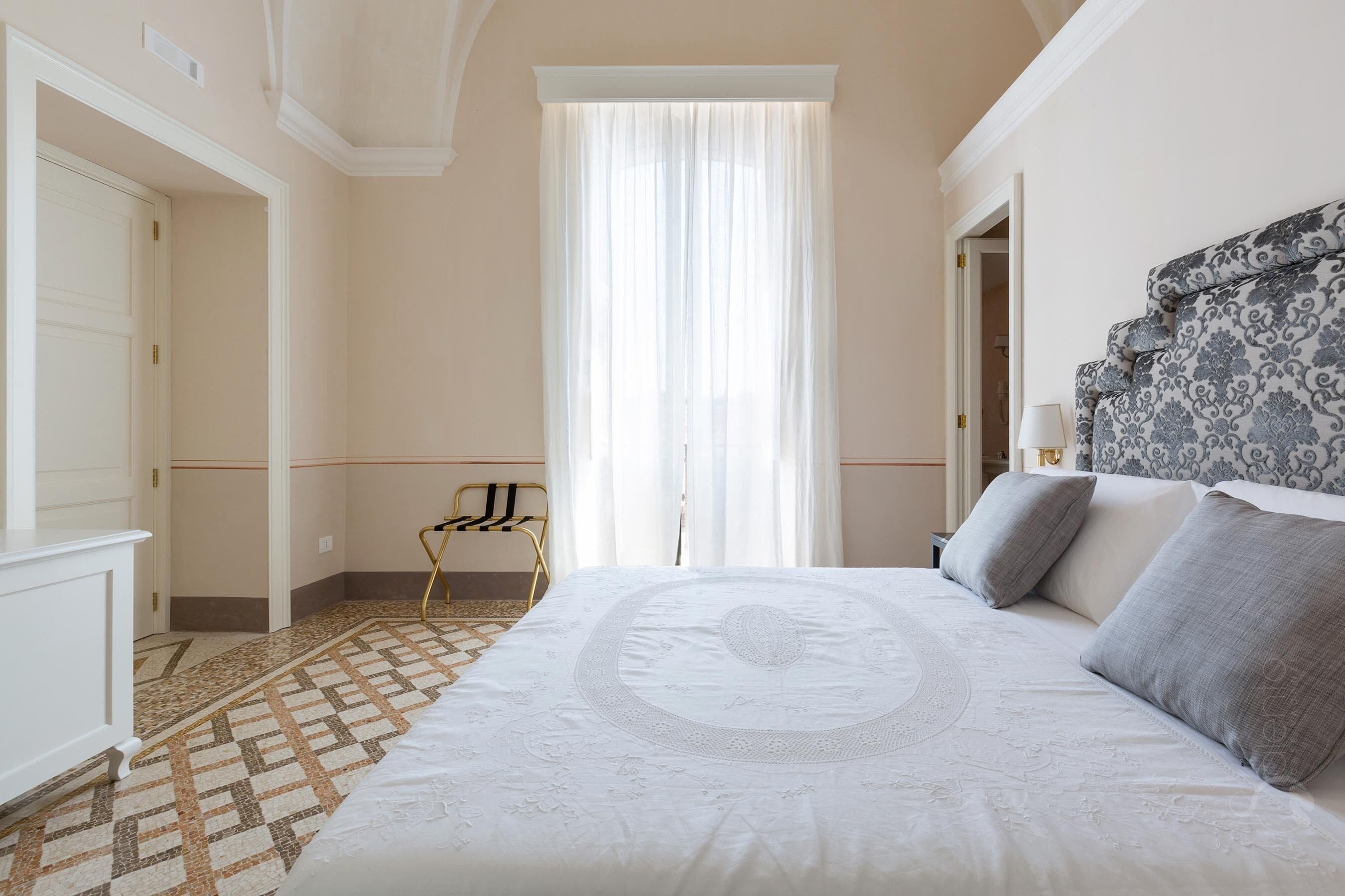 Une chambre à coucher avec un grand lit, une tête de lit à motifs, une chaise et une fenêtre ornée de rideaux légers.