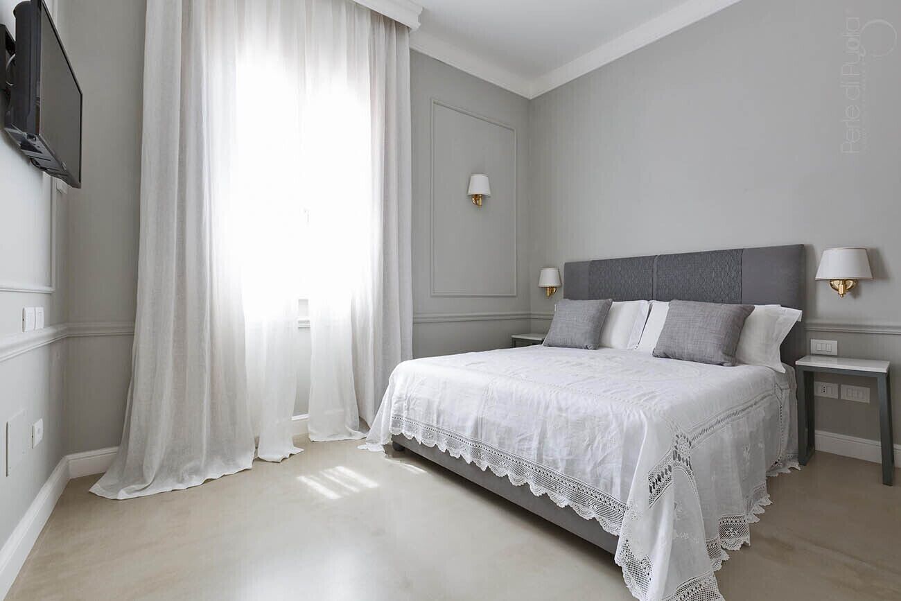 Un dormitorio con cama, dos mesitas de noche, televisor montado en la pared y ventana con cortinas traslúcidas.