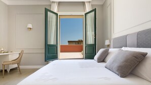 A hotel room with a large bed, a desk, and a chair. The room has a view of a building through an open door. - Palazzo Fasti (Casarano)