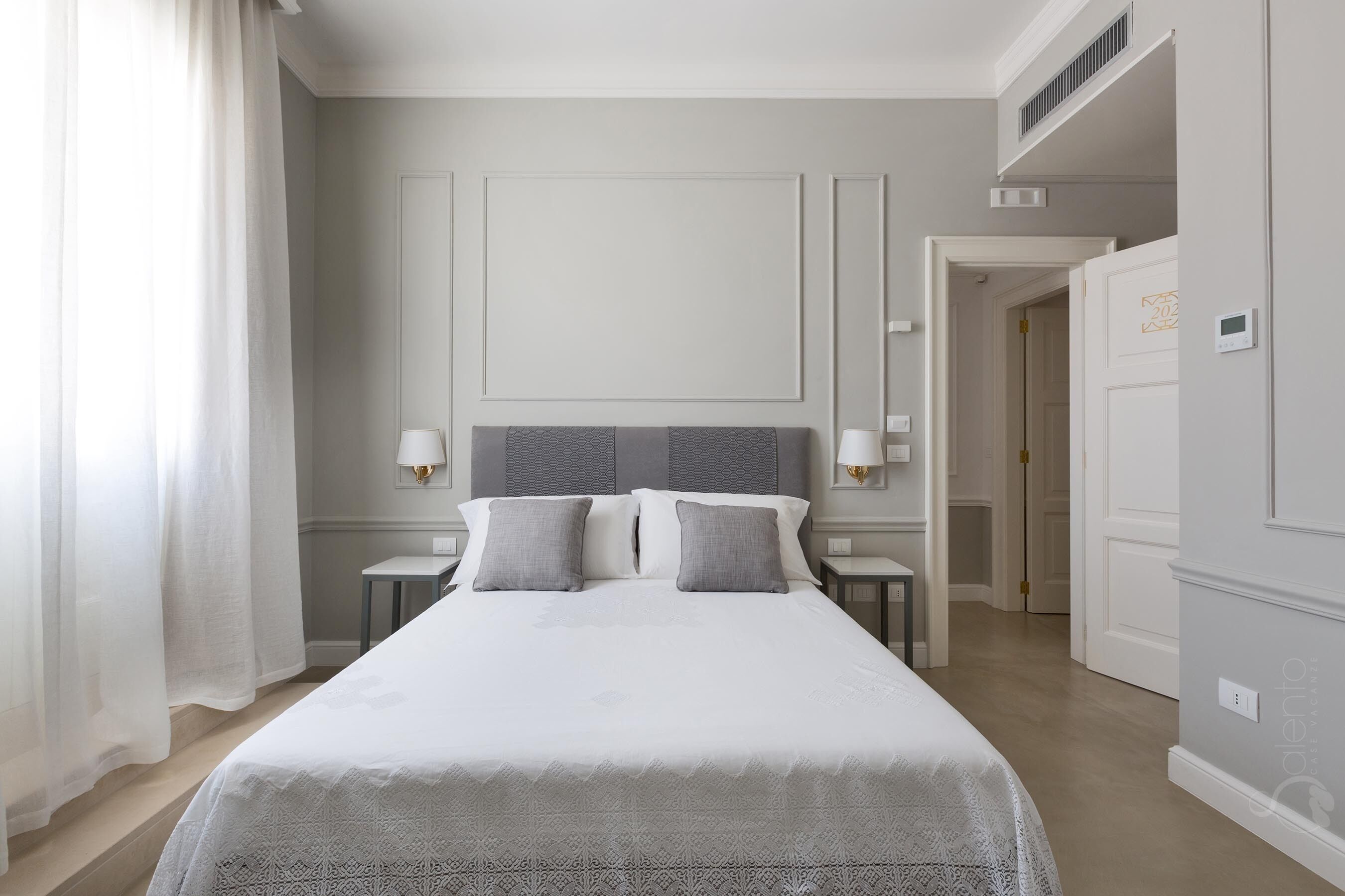 Une chambre d’hôtel avec un grand lit, deux tables de chevet avec des lampes, une porte avec une serrure et un thermostat fixé au mur.
