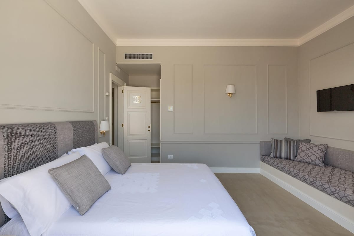 A hotel room with a large bed, a sofa, a TV mounted on the wall, and a door leading to a hallway.