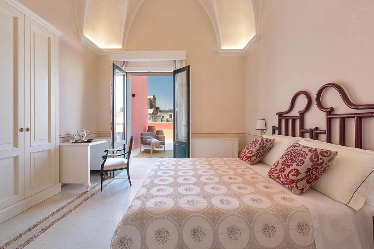 A bedroom with a large bed, a desk, and a chair. There is a view of the outside through an open door.