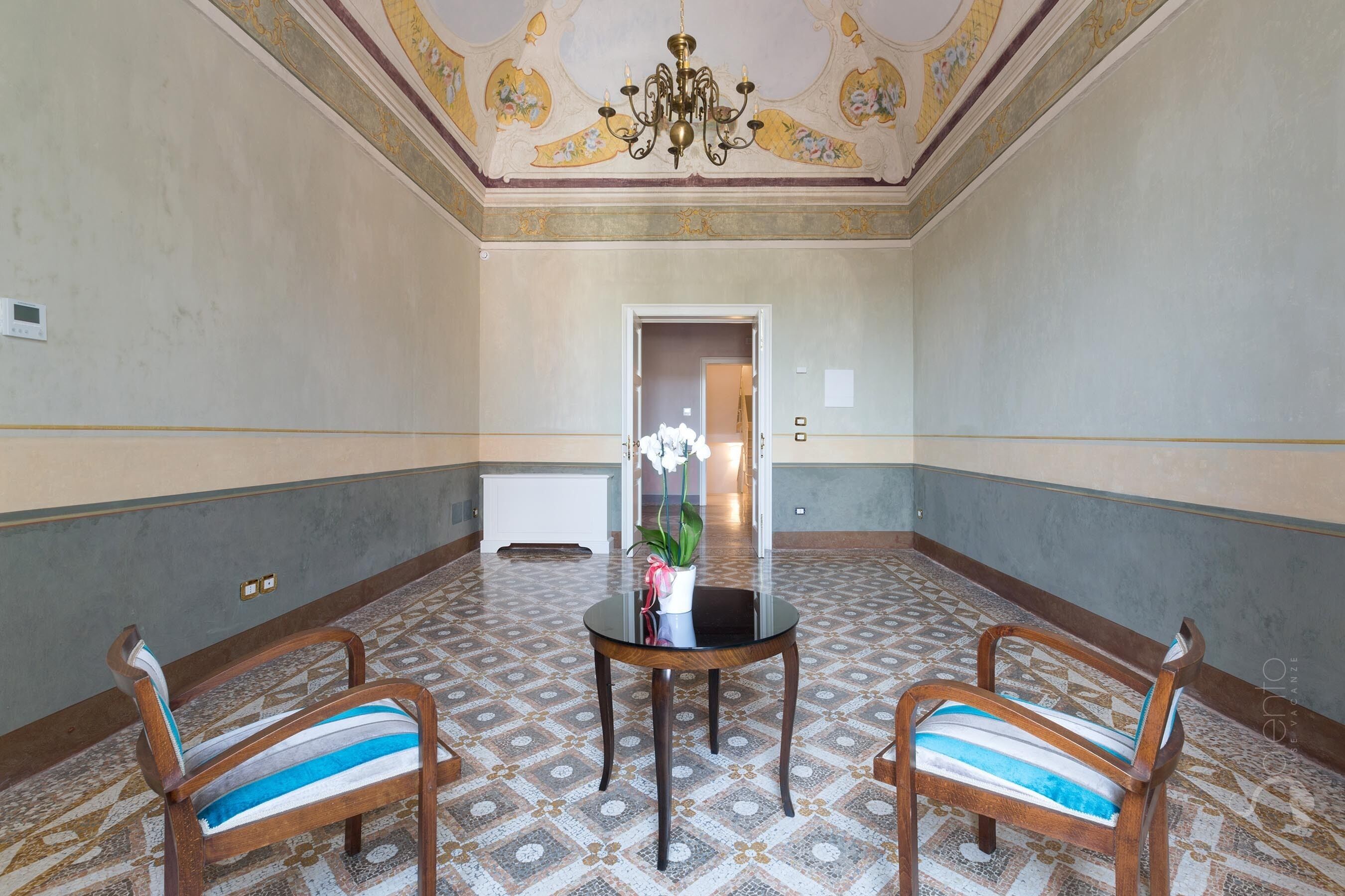 Une pièce dotée d’un sol à motifs, de deux chaises en bois avec des coussins à rayures, d’une table ronde ornée d’un vase de fleurs et d’un plafond décoratif.