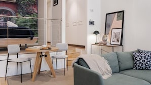 A modern living room with a sofa, a dining table with chairs, a staircase, and a view of a balcony with a person sitting.