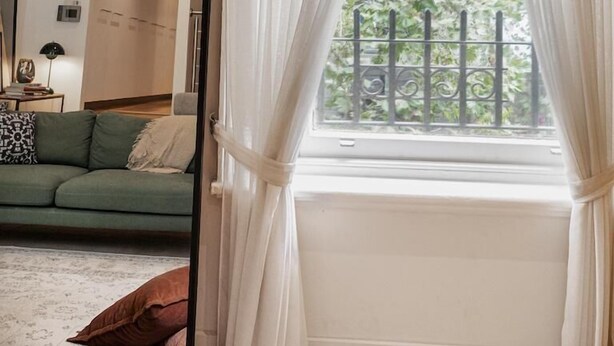 A room with a large window covered by sheer curtains, a green sofa, a mirror, and a view of greenery outside.