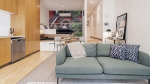 A modern living room with a green sofa, a dining area with a table and chairs, and a kitchen with wooden cabinets and a dishwasher.