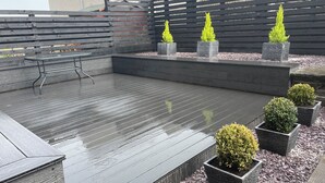 Eine Terrasse mit Holzdeck, einem Metalltisch und -stühlen sowie vier quadratischen Pflanzkübeln mit grünen Sträuchern.
