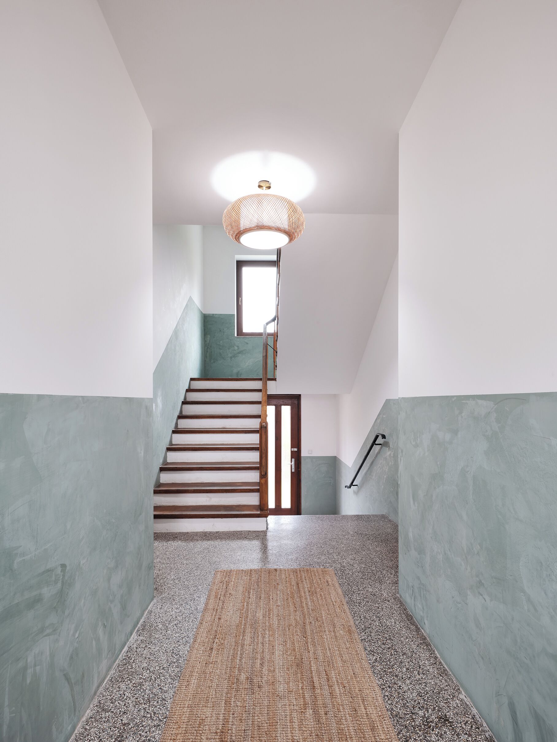 A staircase with a textured wall, a wooden handrail, and a woven light fixture.