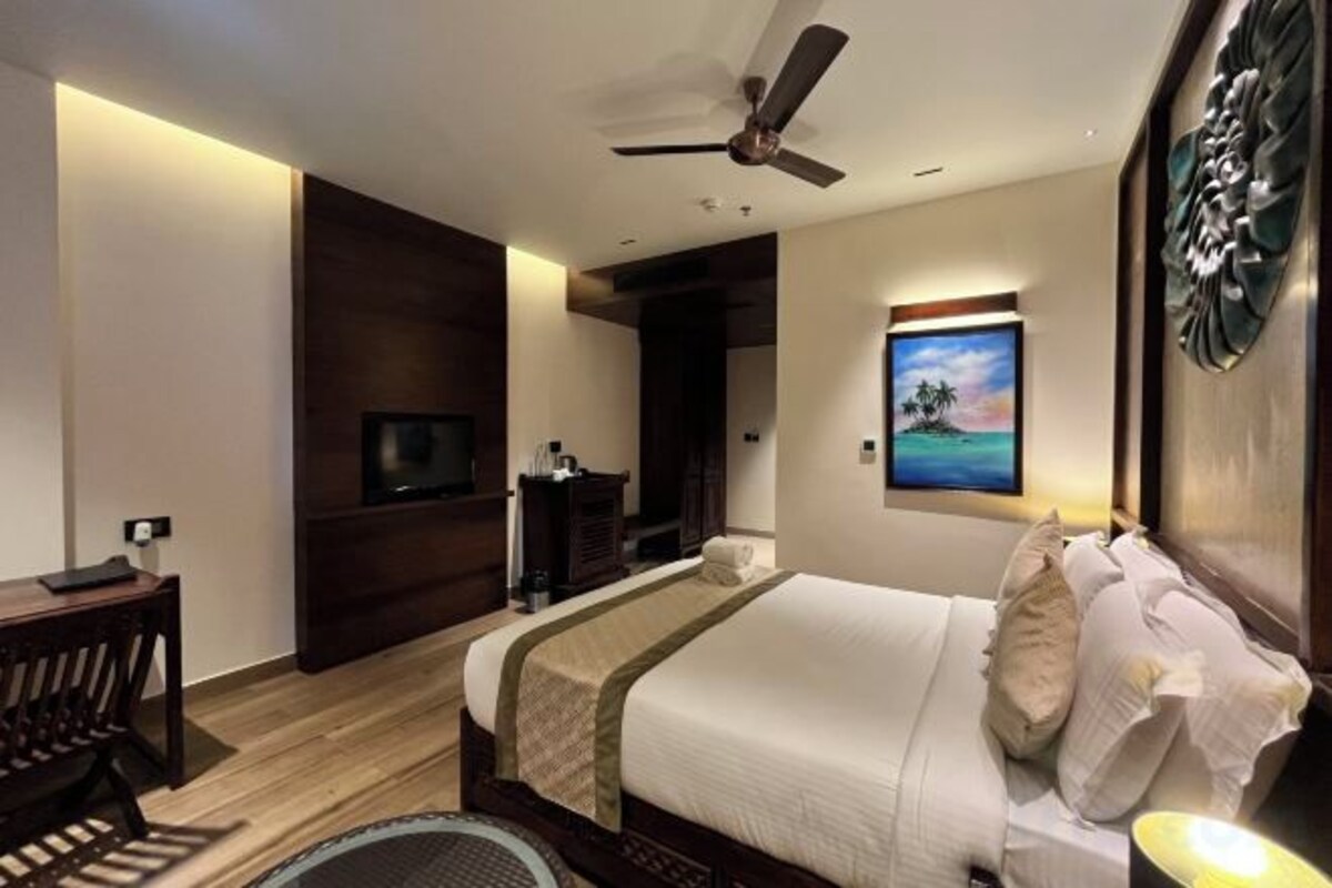 A hotel room with a bed, a desk, a chair, a ceiling fan, a TV, and a painting on the wall.