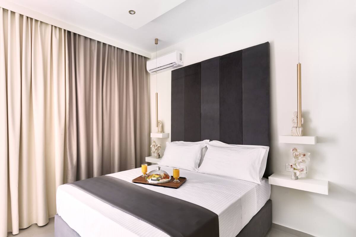 A modern hotel room with a large bed, a bedside table with a fruit bowl, and a wall-mounted air conditioning unit.