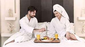 Un couple, vêtu de longues robes blanches, est assis sur un lit et prend son repas sur un plateau garni de nourriture et de boissons.