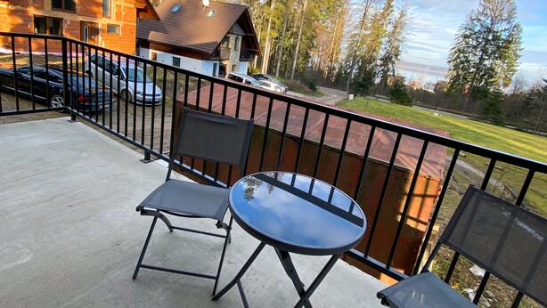 A balcony with two chairs and a table, overlooking a wooden cabin and a forested area.