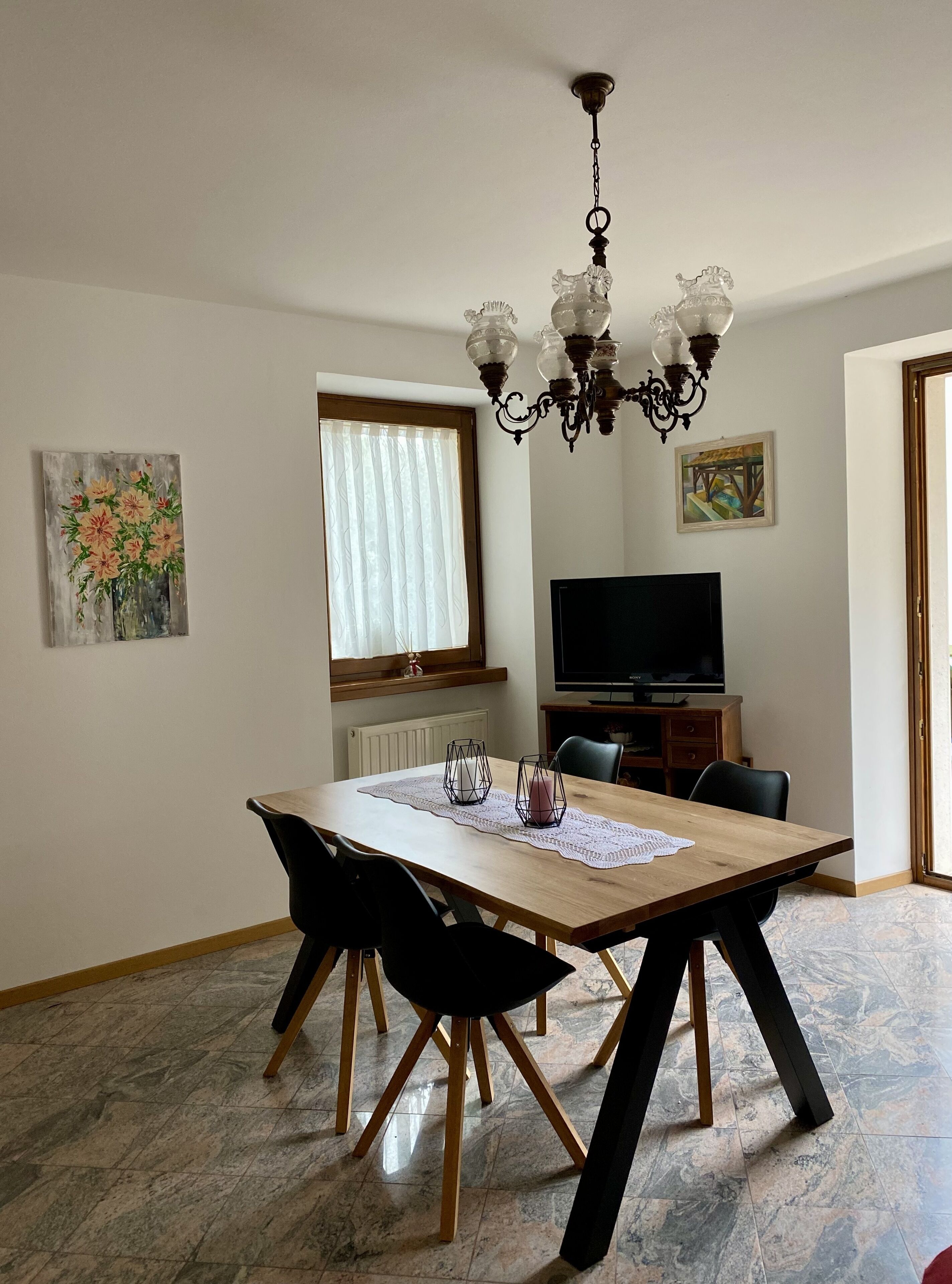Un comedor con mesa de madera, sillas negras, una araña y un televisor al fondo.