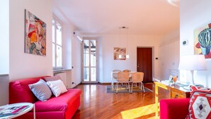 A bright living room with a red sofa, a dining area with wicker chairs, and a kitchen area with a white cabinet and a lamp. - Gerbett Apartment With Terrace by Wonderful Italy (Como)