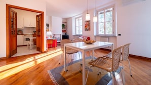 A spacious dining area with a white table and wicker chairs, a kitchen in the background, and large windows allowing natural light. - Gerbett Apartment With Terrace by Wonderful Italy (Como)