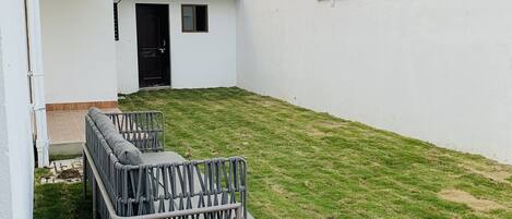 Un espacio al aire libre pequeño y cercado con césped, una banca de metal y una puerta que da a un edificio.