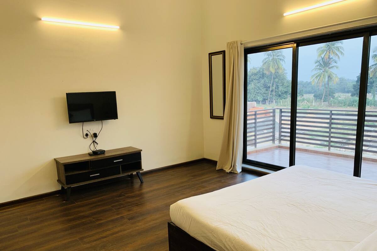 A bedroom with a large bed, a TV mounted on the wall, a wooden dresser, and a balcony with a view of palm trees.