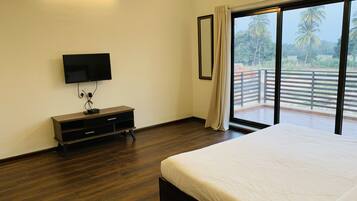 A bedroom with a large bed, a TV mounted on the wall, a wooden dresser, and a balcony with a view of palm trees.