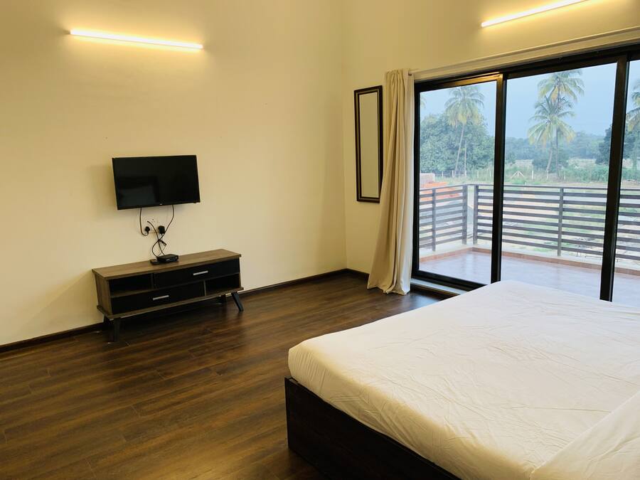 A bedroom with a large bed, a TV mounted on the wall, a wooden dresser, and a balcony with a view of palm trees.
