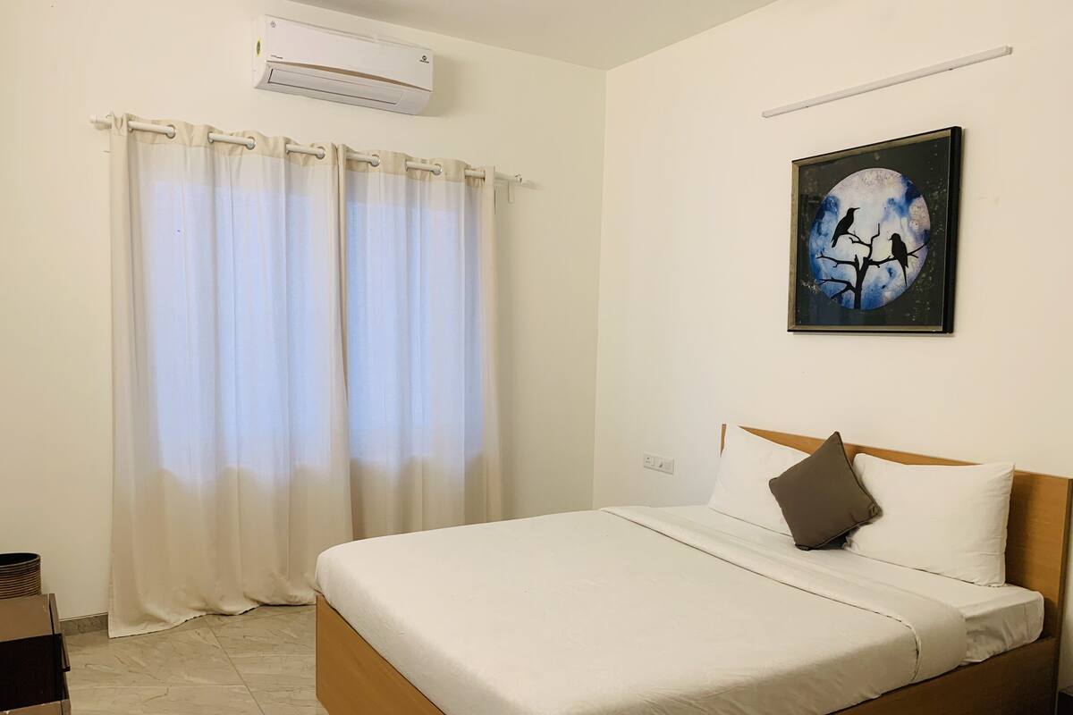 A neatly made bed with white bedding and a brown pillow, a wall-mounted air conditioner, and a painting of birds on a tree branch.