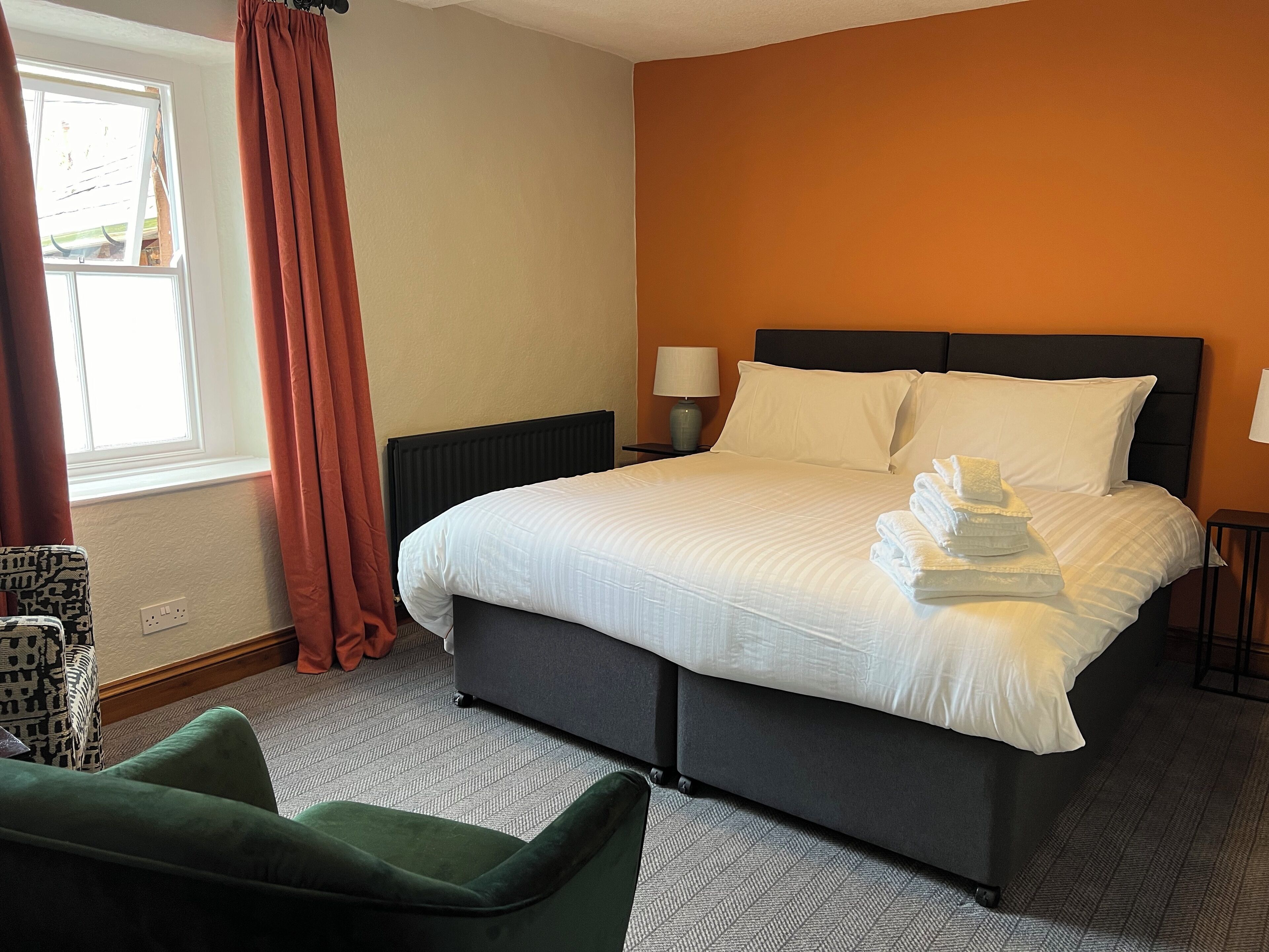 Een hotelkamer met een groot bed, twee kussens, een nachtkastje met een lamp, een stoel en een raam met gordijnen.