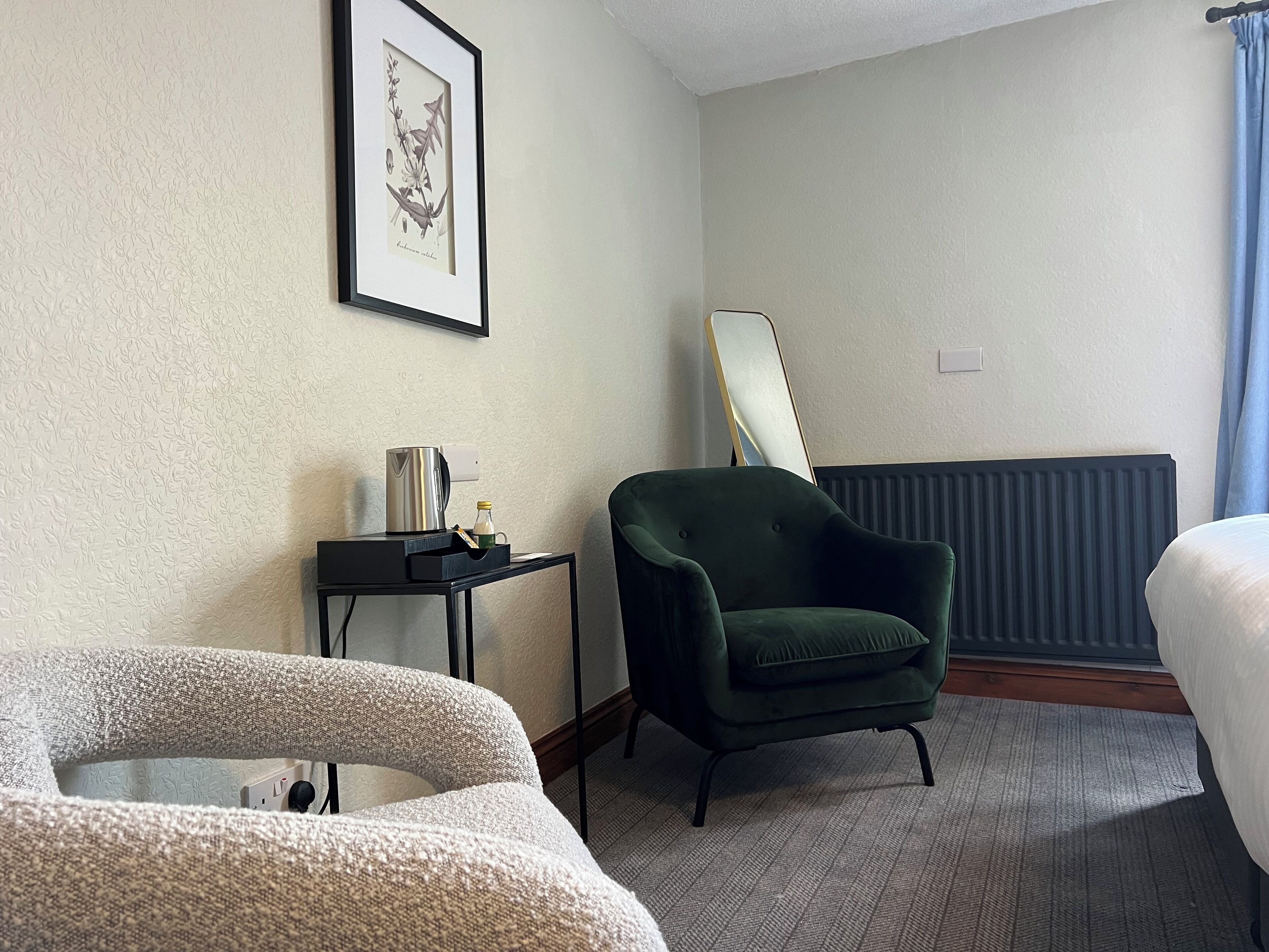 Habitación de hotel con cama, un sillón verde, una mesita con una tetera y una botella, un grabado botánico enmarcado en la pared y un radiador.