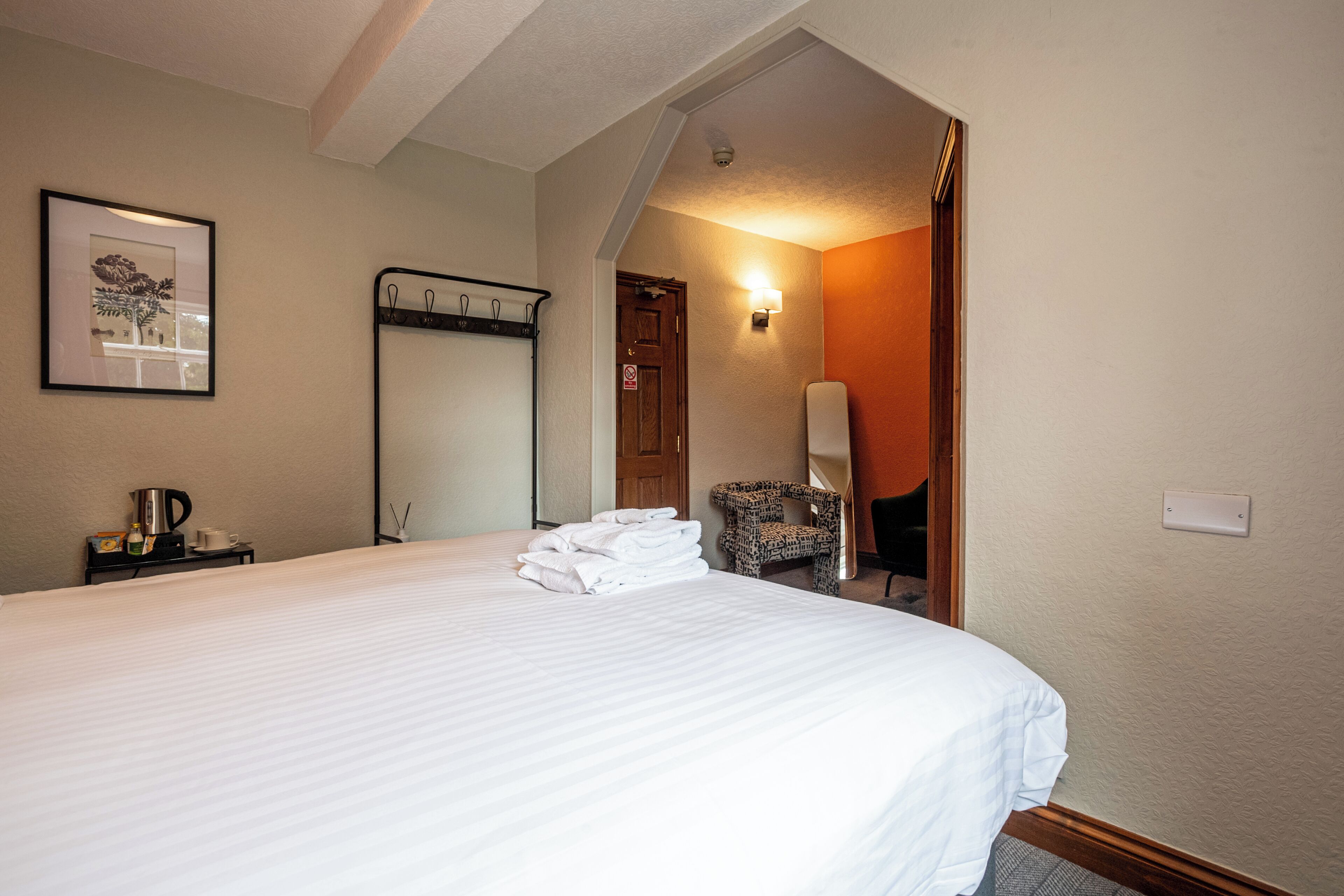 Hotelkamer met een groot bed, een stoel, een kapstok, een ingelijst schilderij en een deur naar een andere kamer.