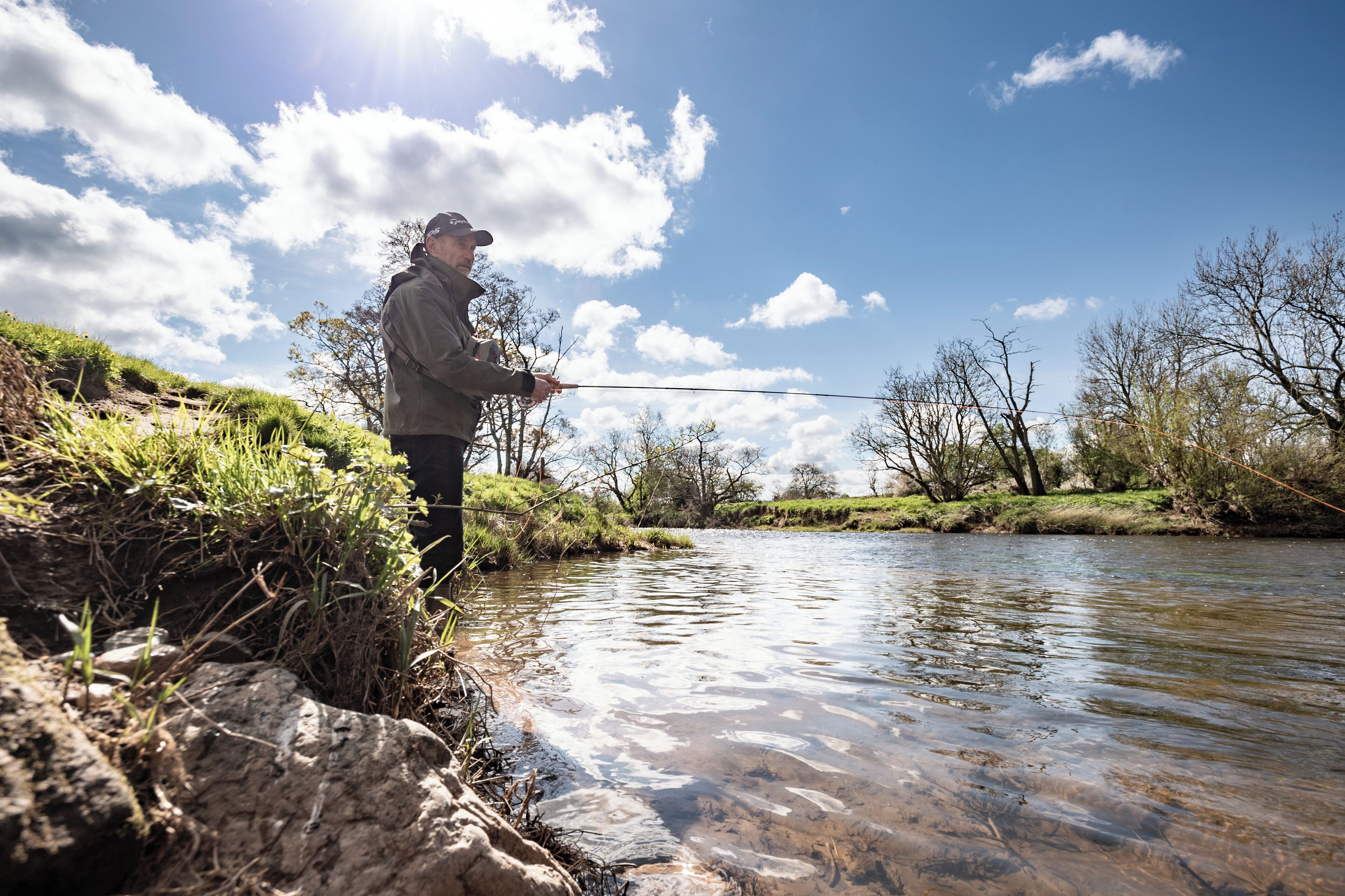 A person fishing by a clear stream with a rocky bank and sparse vegetation.