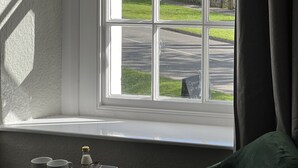 A room with a window overlooking a street, a green chair, a small table with cups and a bottle, and a sign indicating the room's opening hours. - The Kings Arms (Penrith)