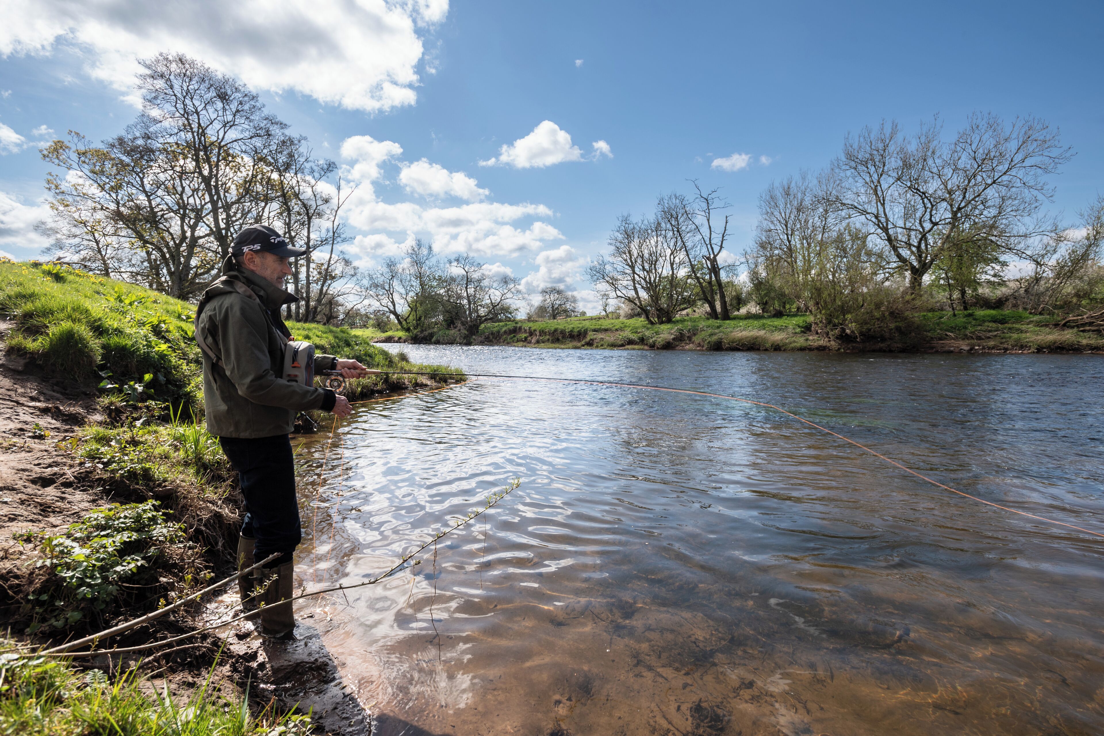 A person standing in shallow water near a riverbank, holding a fishing rod.