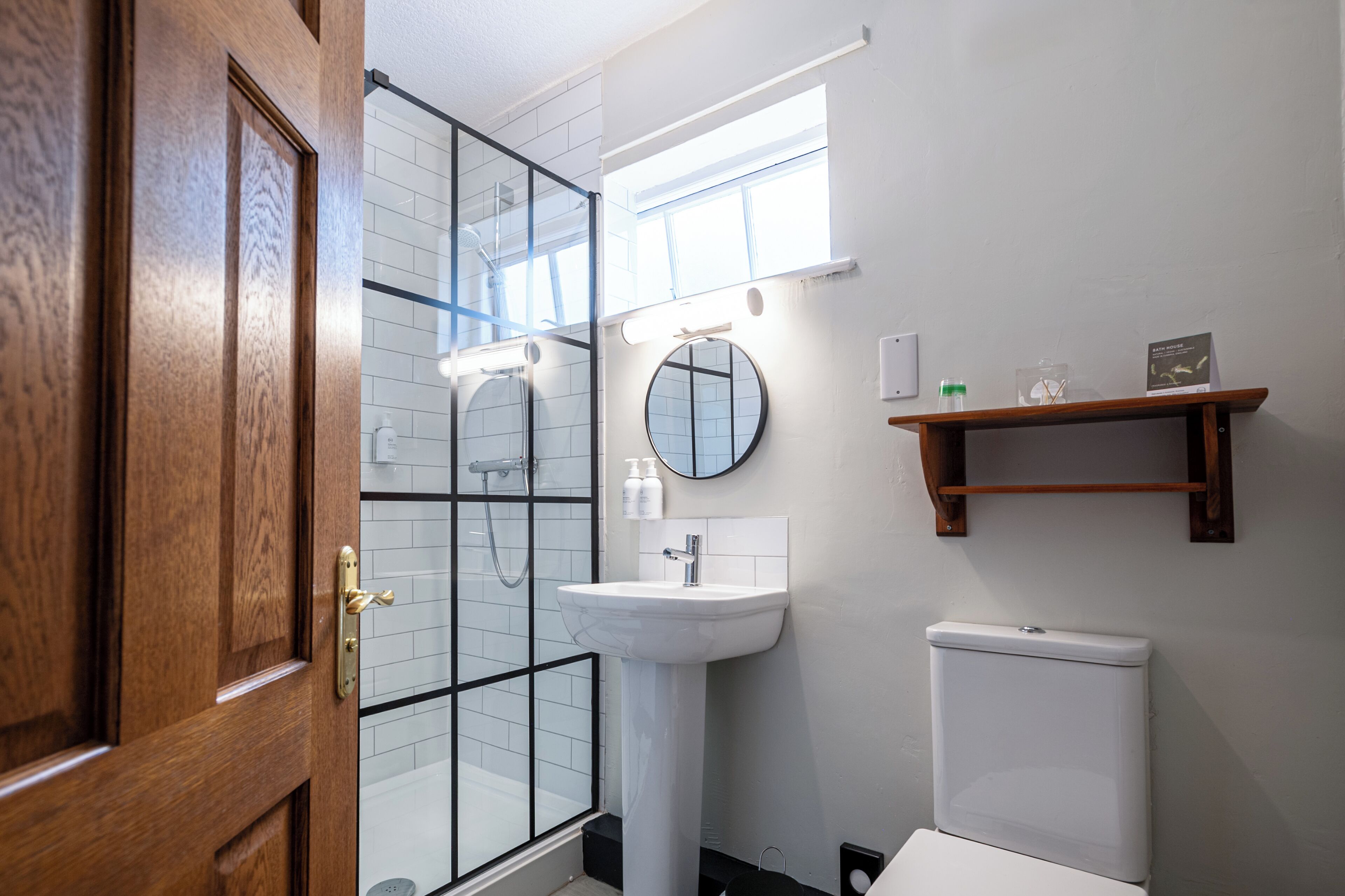 Un baño con mampara de vidrio, un lavamanos blanco, inodoro y repisa de madera con artículos de aseo.