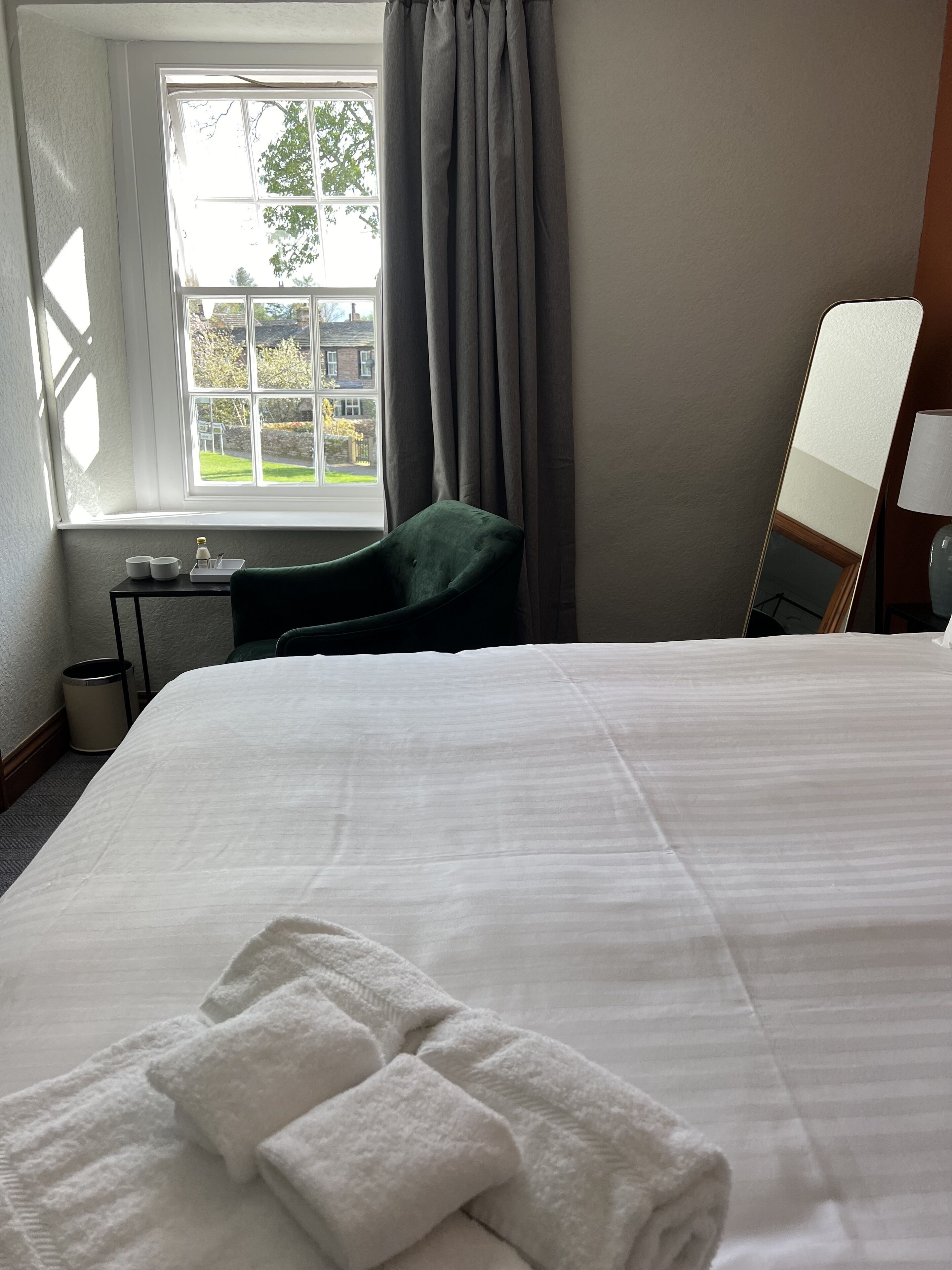 Habitación de hotel con una cama grande, una silla, una mesita con tazas y un ventanal con cortinas.