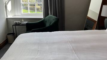 Habitación de hotel con una cama grande, una silla, una mesita con tazas y un ventanal con cortinas.