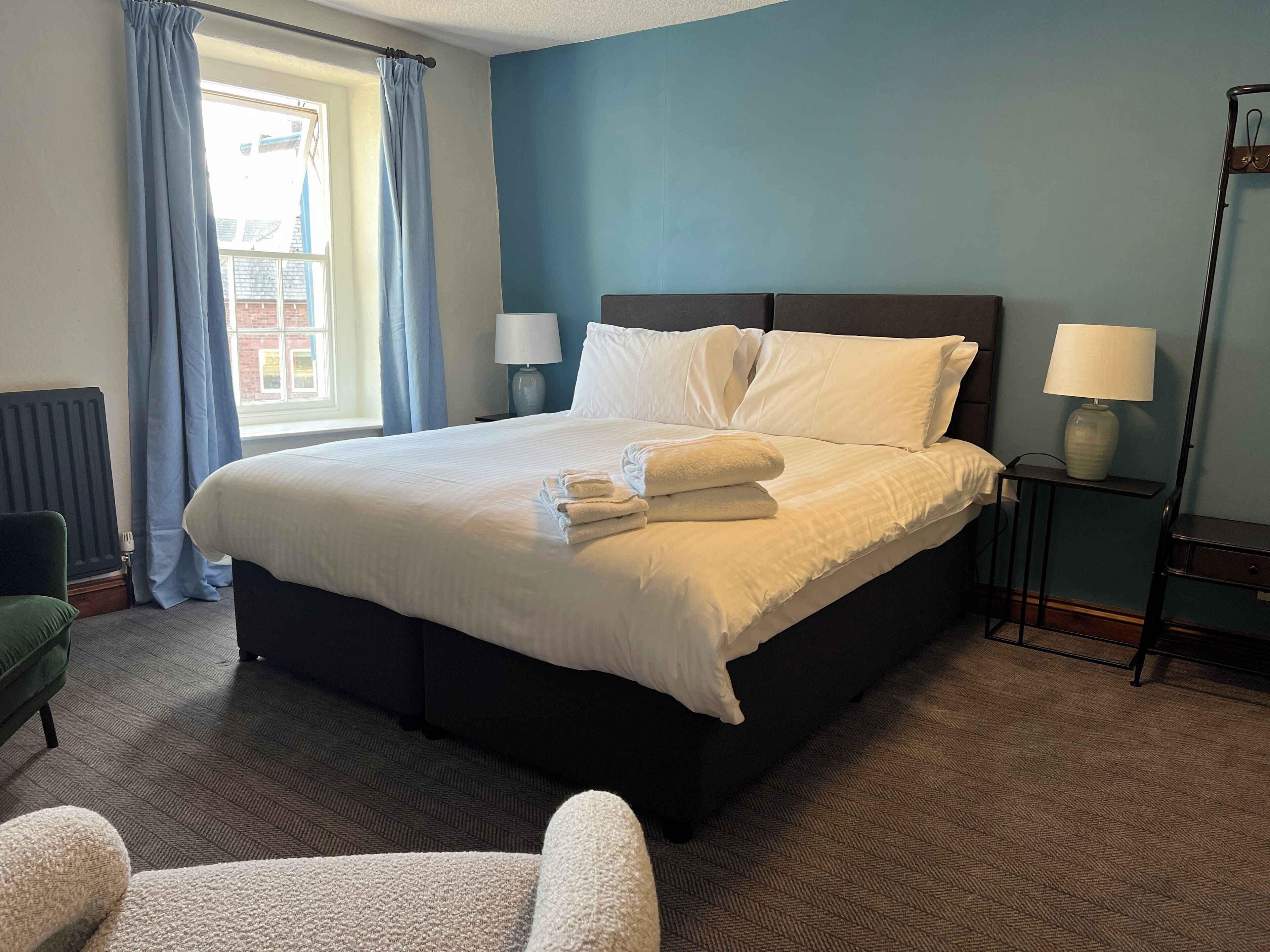 A neatly made bed with white linens and folded towels, a bedside lamp, and a view of a building through a window with blue curtains.