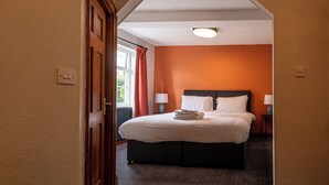 A hotel room with a large bed, two bedside tables with lamps, an orange wall, and a window with red curtains. - The Kings Arms (Penrith)
