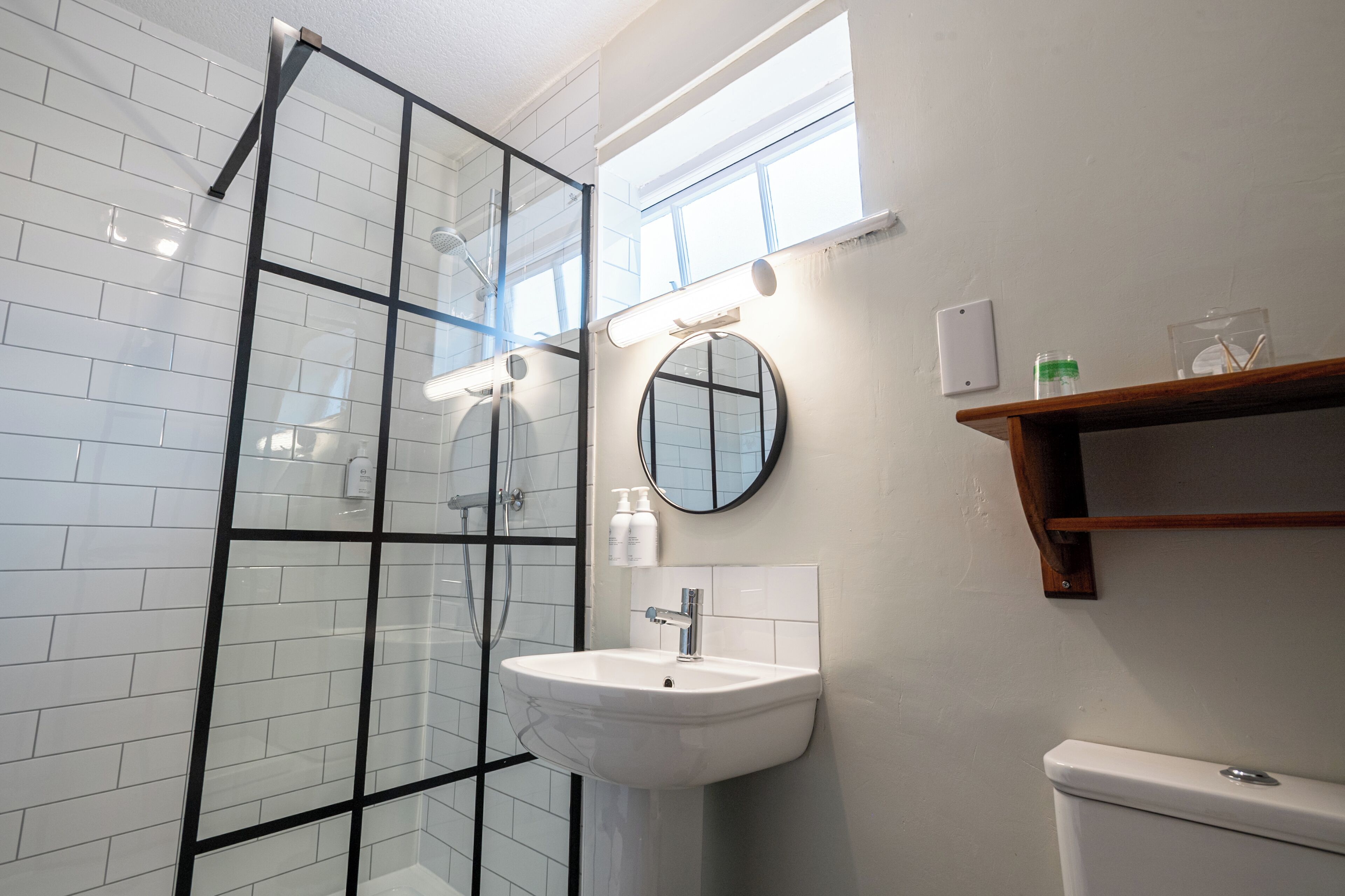 A modern bathroom with a glass shower enclosure, a white sink with a mirror, and a wooden shelf above the toilet.