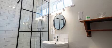 A modern bathroom with a glass shower enclosure, a white sink with a mirror, and a wooden shelf above the toilet.