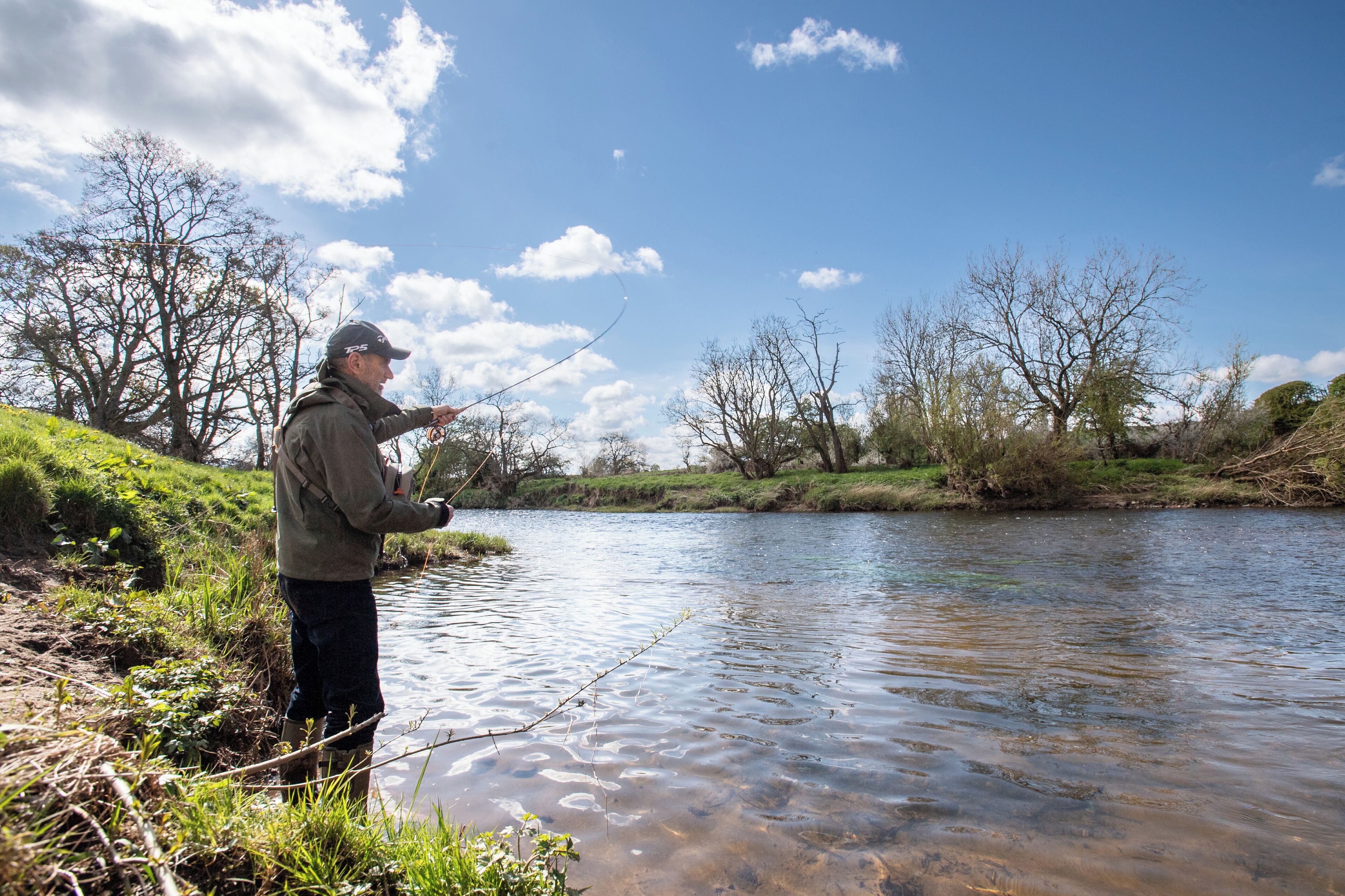 A person fishing by a calm river with trees and grassy banks.