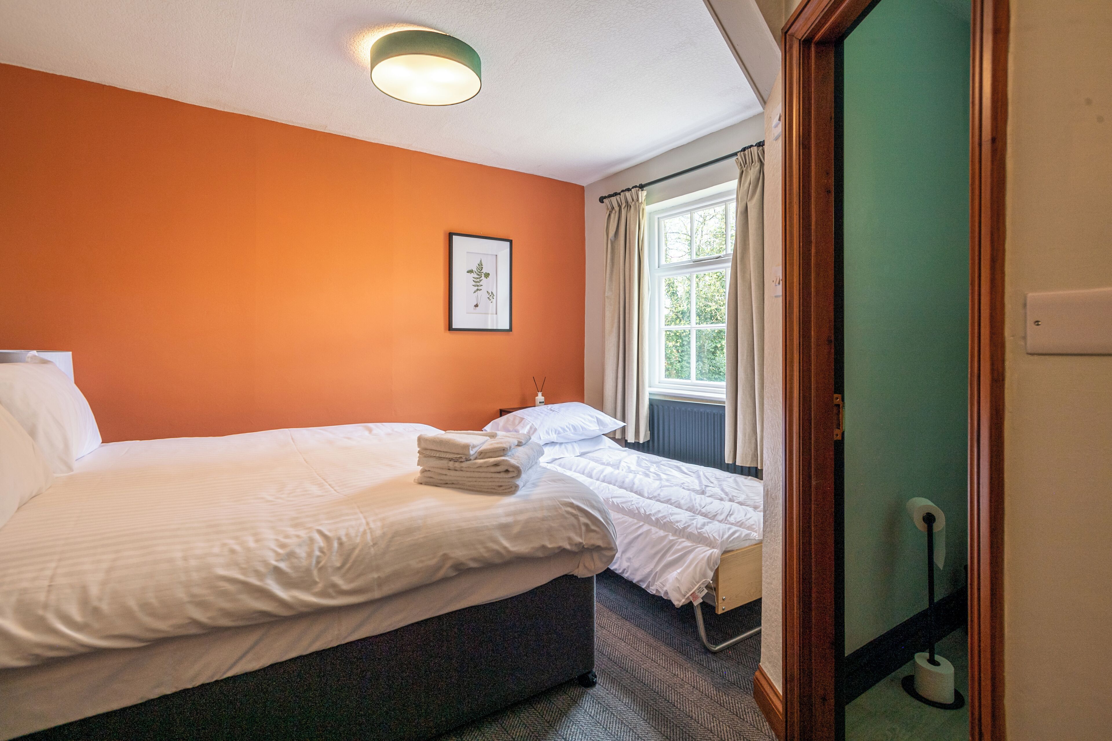 A hotel room with two beds, a framed picture on the wall, a window with curtains, and a radiator.