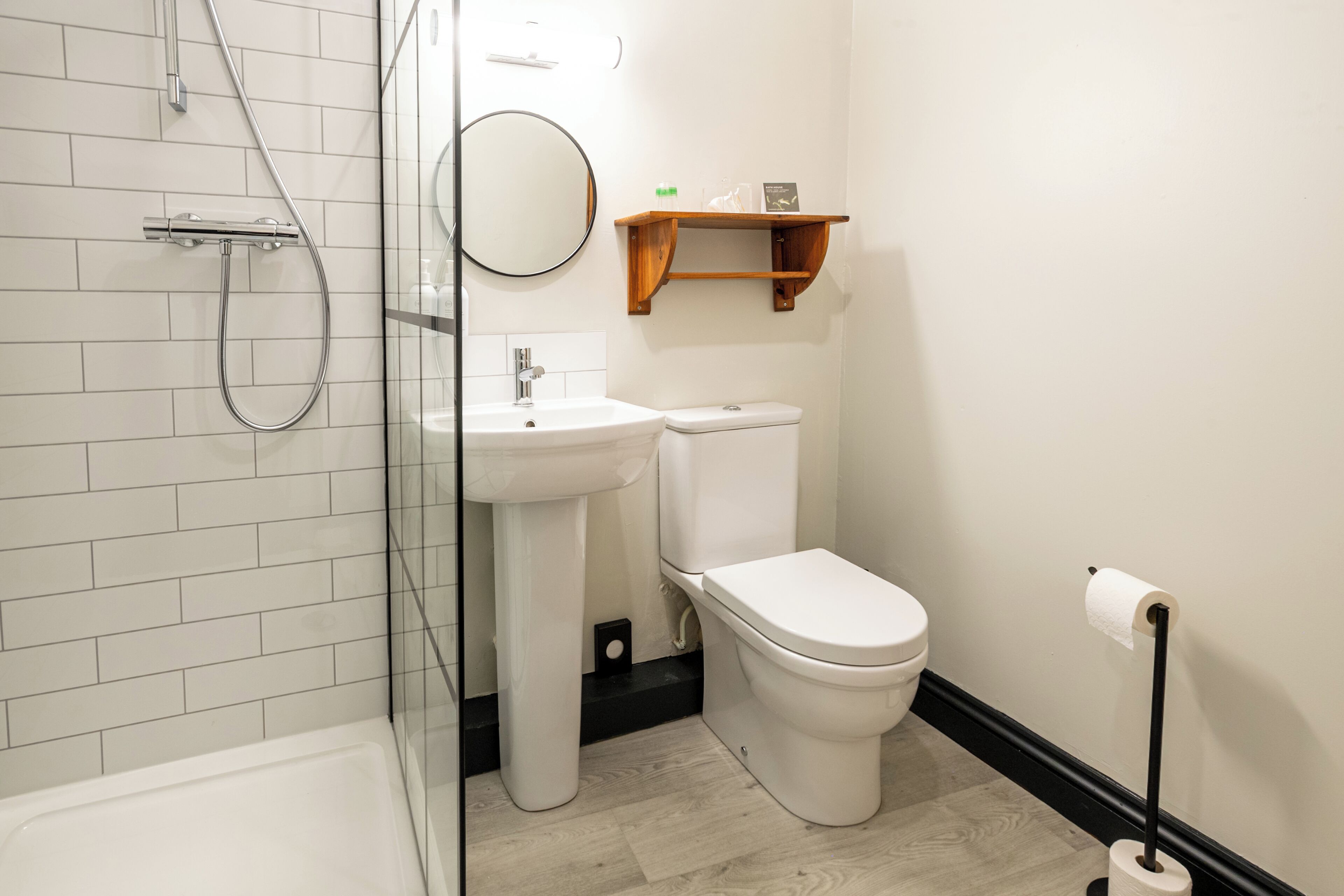 A modern bathroom with a glass shower enclosure, a white sink, a toilet, and a wooden shelf above the toilet.