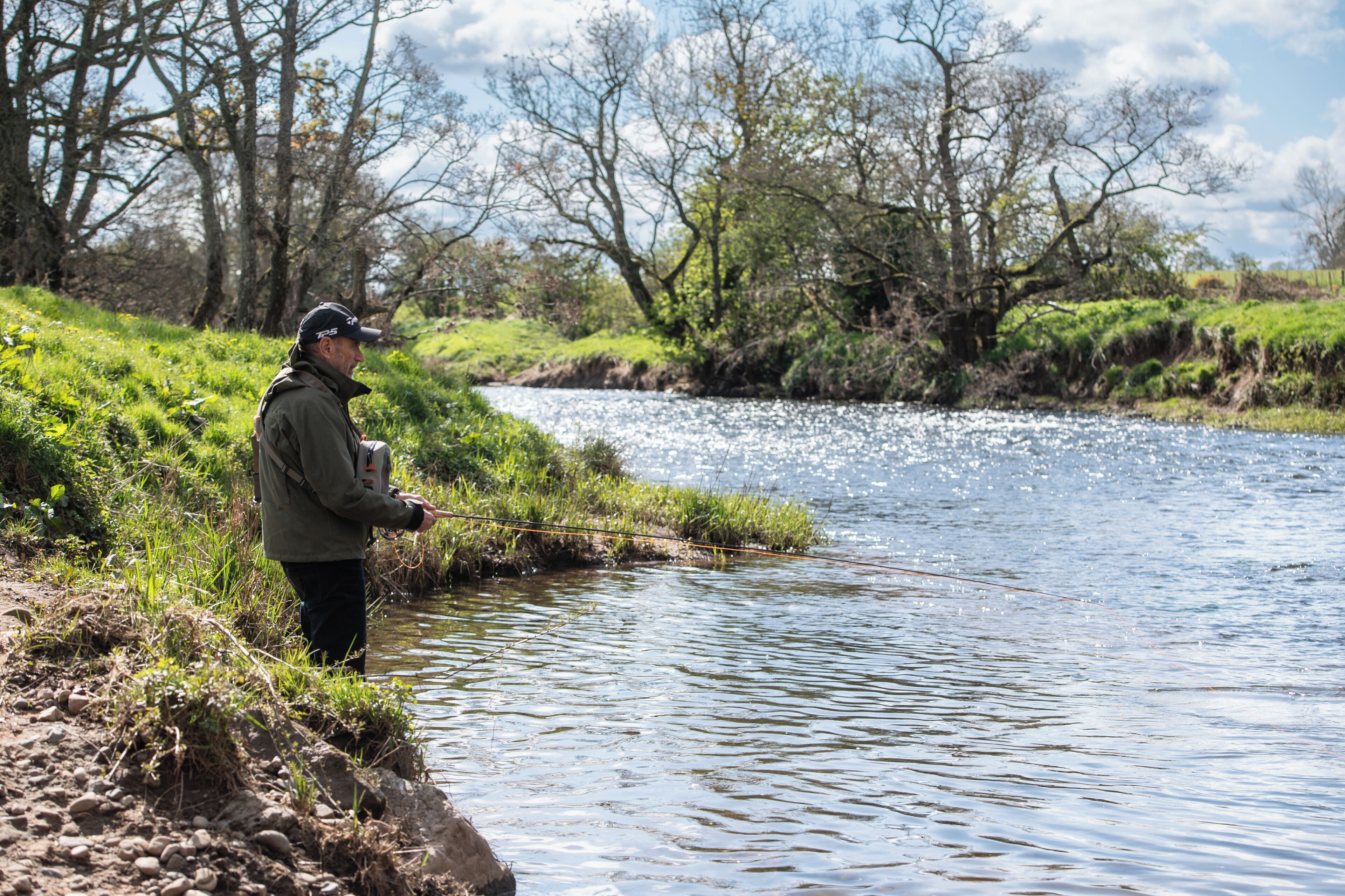 A person standing by a river with a fishing rod, surrounded by greenery and trees.