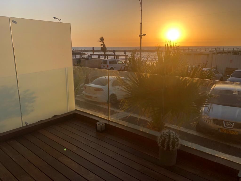 A balcony with a view of the sunset over the ocean, a parked car, and a potted plant.