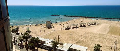 A beach with clear blue water, a sandy shore, and a few structures including a building with a sign and a small pavilion.