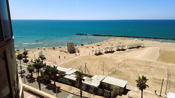 A beach with clear blue water, a sandy shore, and a few structures including a building with a sign and a small pavilion. - LBA Haifa (Haifa)