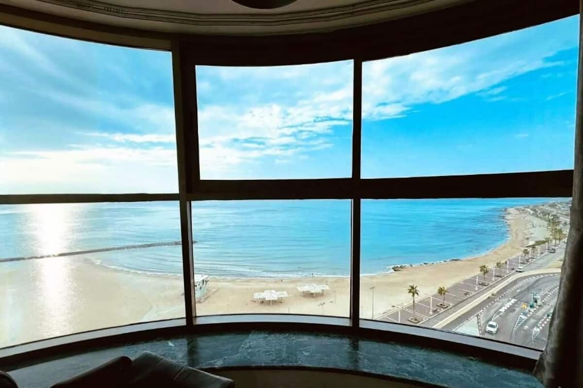 Vista panorámica desde una habitación con ventanas amplias que dan a una playa y al océano.