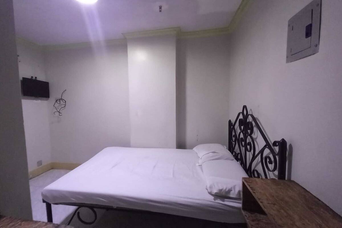 A small, simple room with a single bed, a bedside table, and a wall-mounted light fixture.