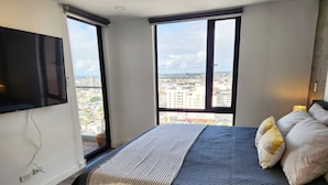 A modern bedroom with a large bed, a TV mounted on the wall, and a city view through large windows.
