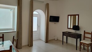 Ein Zimmer mit einem Esstisch, Stühlen, einem Fernseher, einem Schreibtisch mit Stuhl, einer Lampe und einem Spiegel.